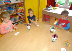 Troje dzieci siedzi przy stoliku w ręku trzymają kredki i układają je na stole.